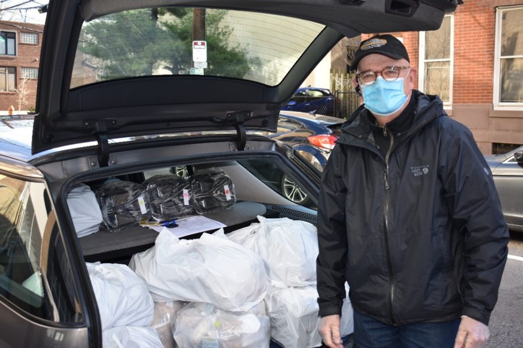 an older man, a volunteer driver in a MANNA hat, disposable face mask, brown glasses, and black windbreaker, stands in front of his open trunk, which is filled with bagged MANNA meals