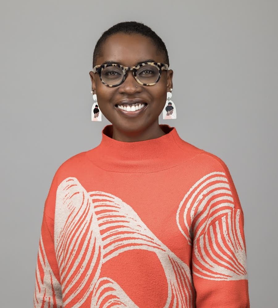 Young Black woman with close-cropped hair, chunky white earrings, thick-rimmed glasses, wearing an orange sweater, smiling
